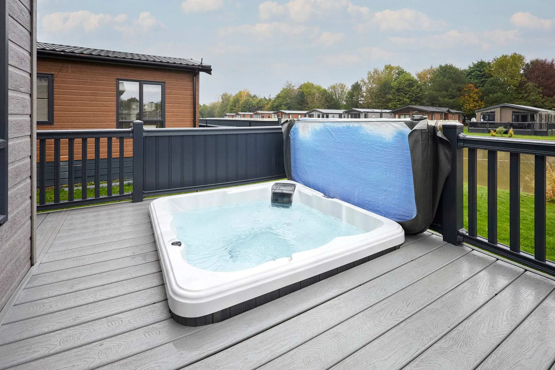 A hot tub sits on a deck beside a railing, with trees and buildings beyond.
