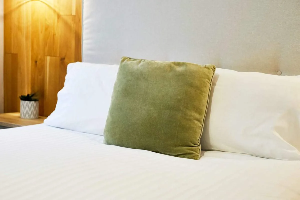 Cama de dormitorio con ropa de cama blanca y un cojín verde.