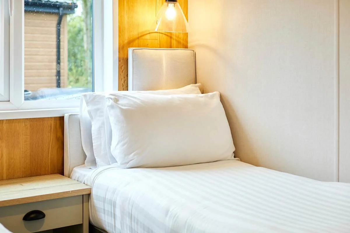 Una cama individual con ropa de cama blanca está junto a una ventana y una lámpara de pared.