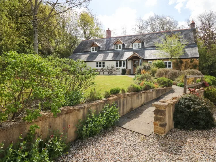 A large country house stands beyond a garden path, surrounded by trees and planted beds.
