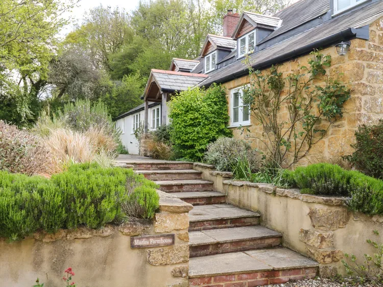 A stone cottage sits beside landscaped garden steps and leafy shrubs.