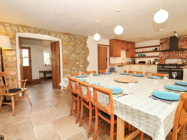 Una gran mesa de comedor ocupa una cocina rústica con armarios de madera y paredes de piedra.