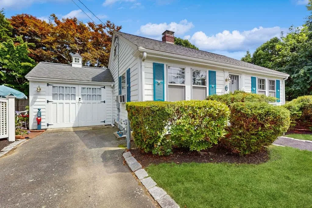 A white house with blue shutters stands behind shrubs beside a paved driveway.