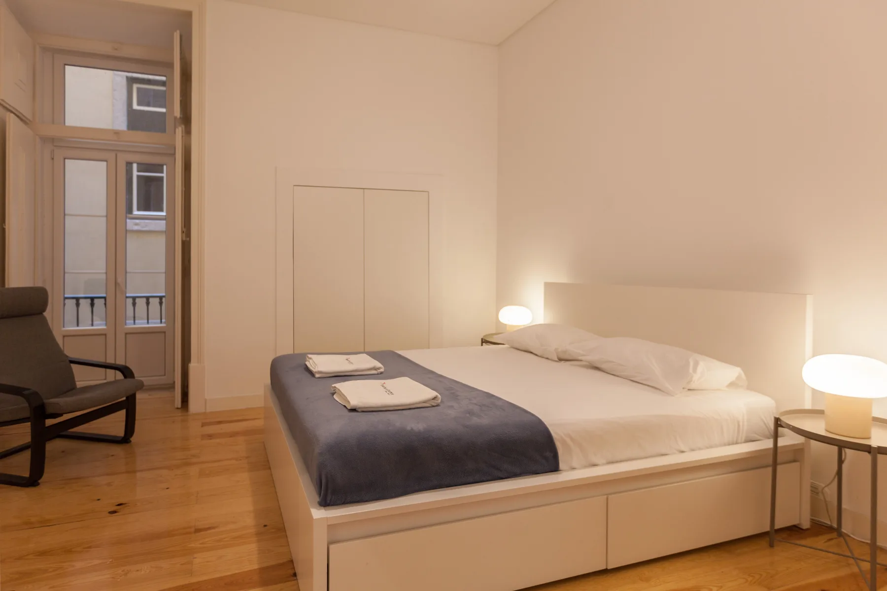Un dormitorio con una cama bien tendida y una silla junto a ventanas altas.