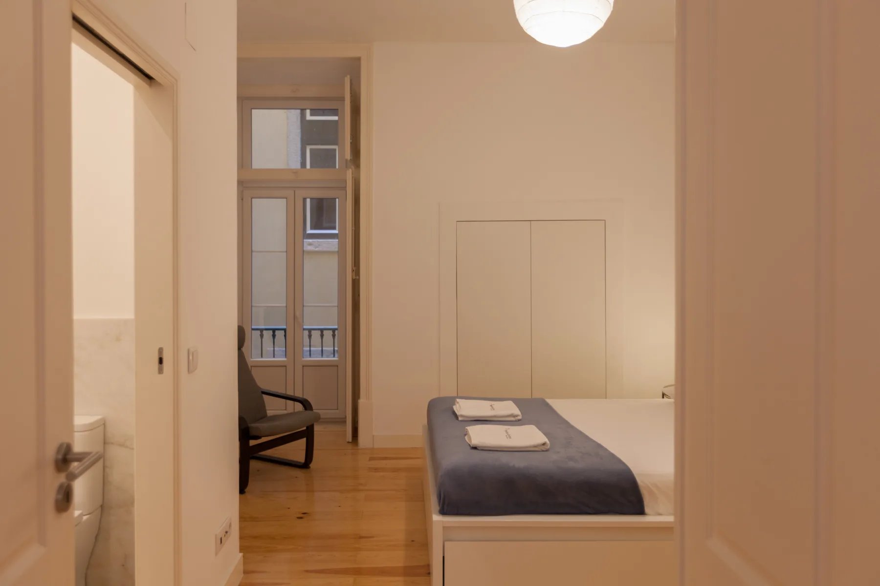Un dormitorio con una cama hecha y una silla junto a ventanas altas.