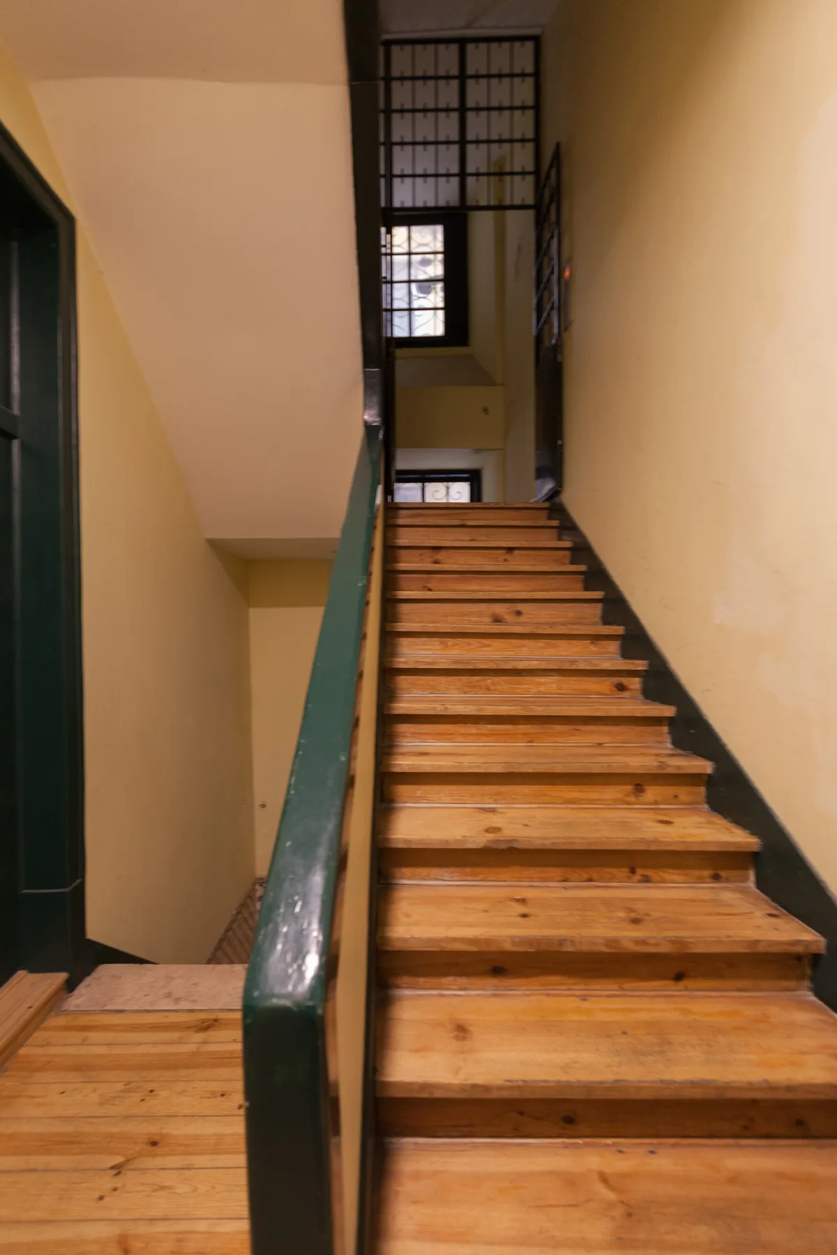 Una escalera de madera sube entre paredes color crema junto a una barandilla verde oscura.