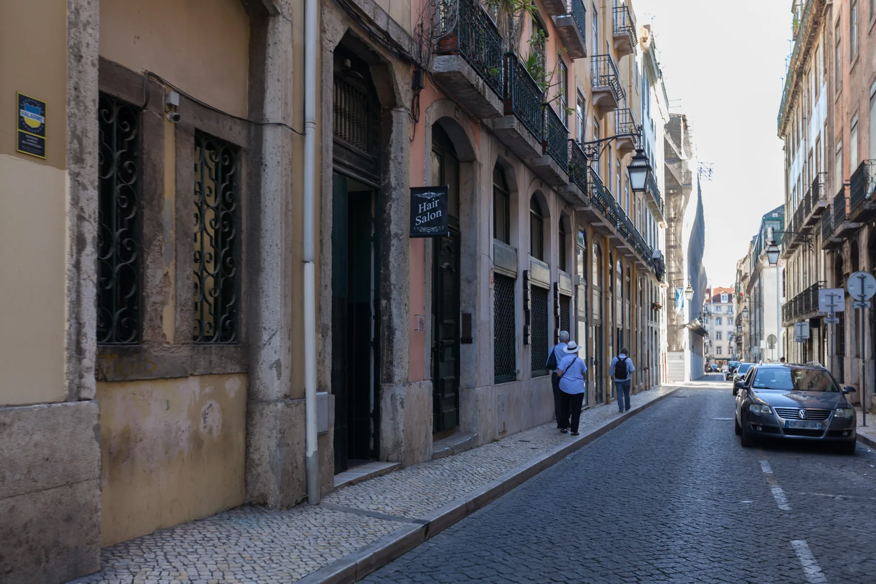 Una calle estrecha empedrada pasa entre edificios altos, con peatones y un coche estacionado.