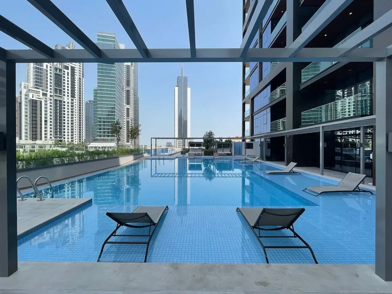 Piscina al aire libre junto a edificios modernos de gran altura, con tumbonas en la terraza.