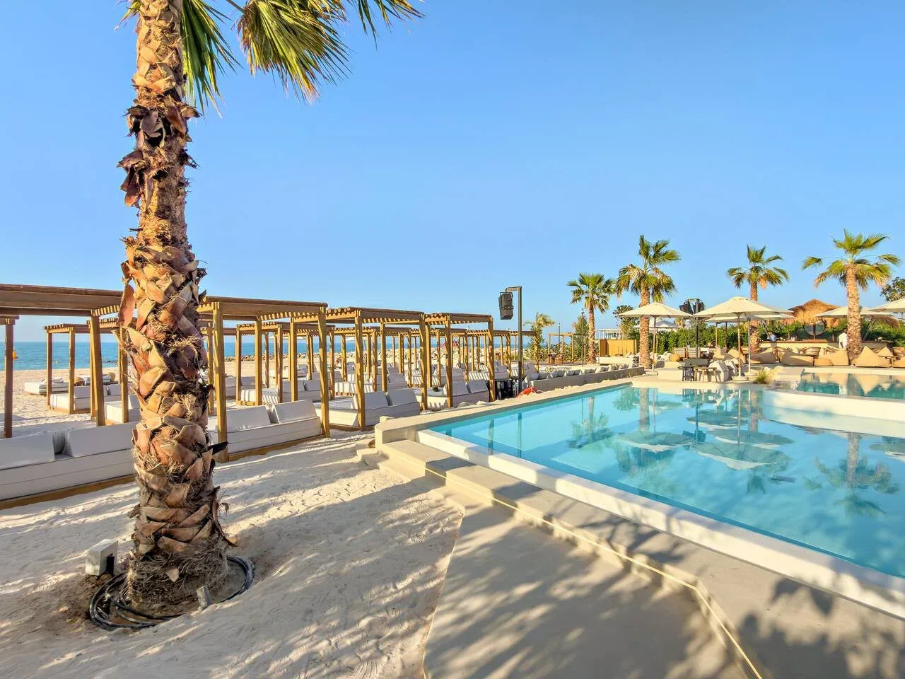Una piscina junto a una playa de arena, con palmeras y asientos a la sombra.