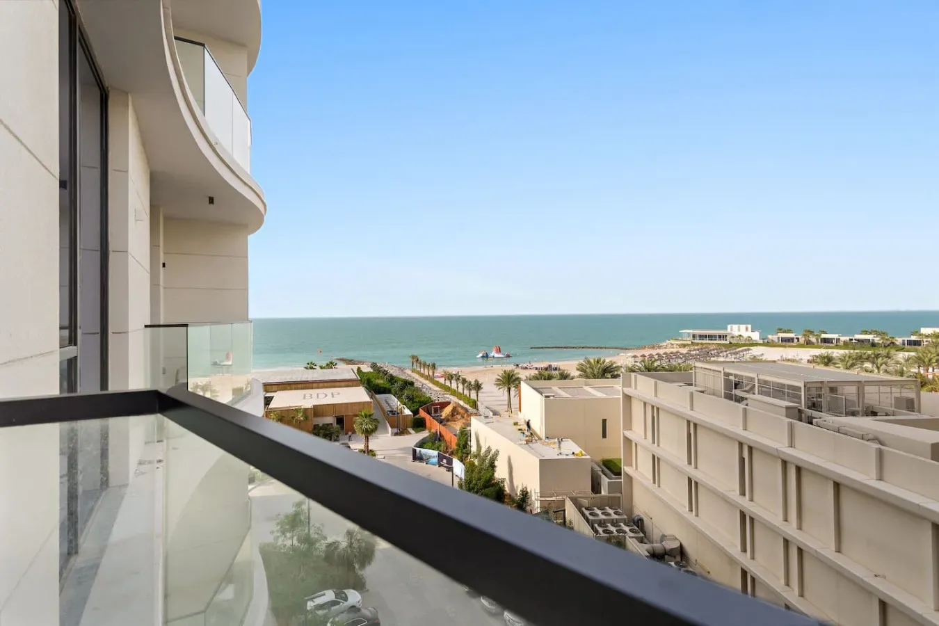 Un balcón da a edificios junto a la playa, palmeras y el mar bajo un cielo despejado.