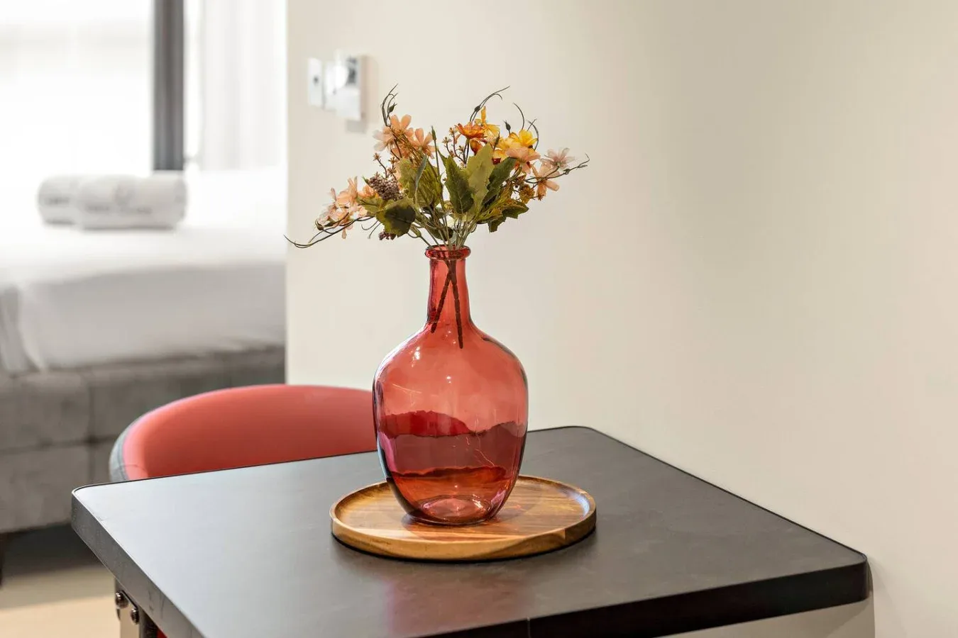 Una mesa con un jarrón de vidrio con flores está junto a una cama.
