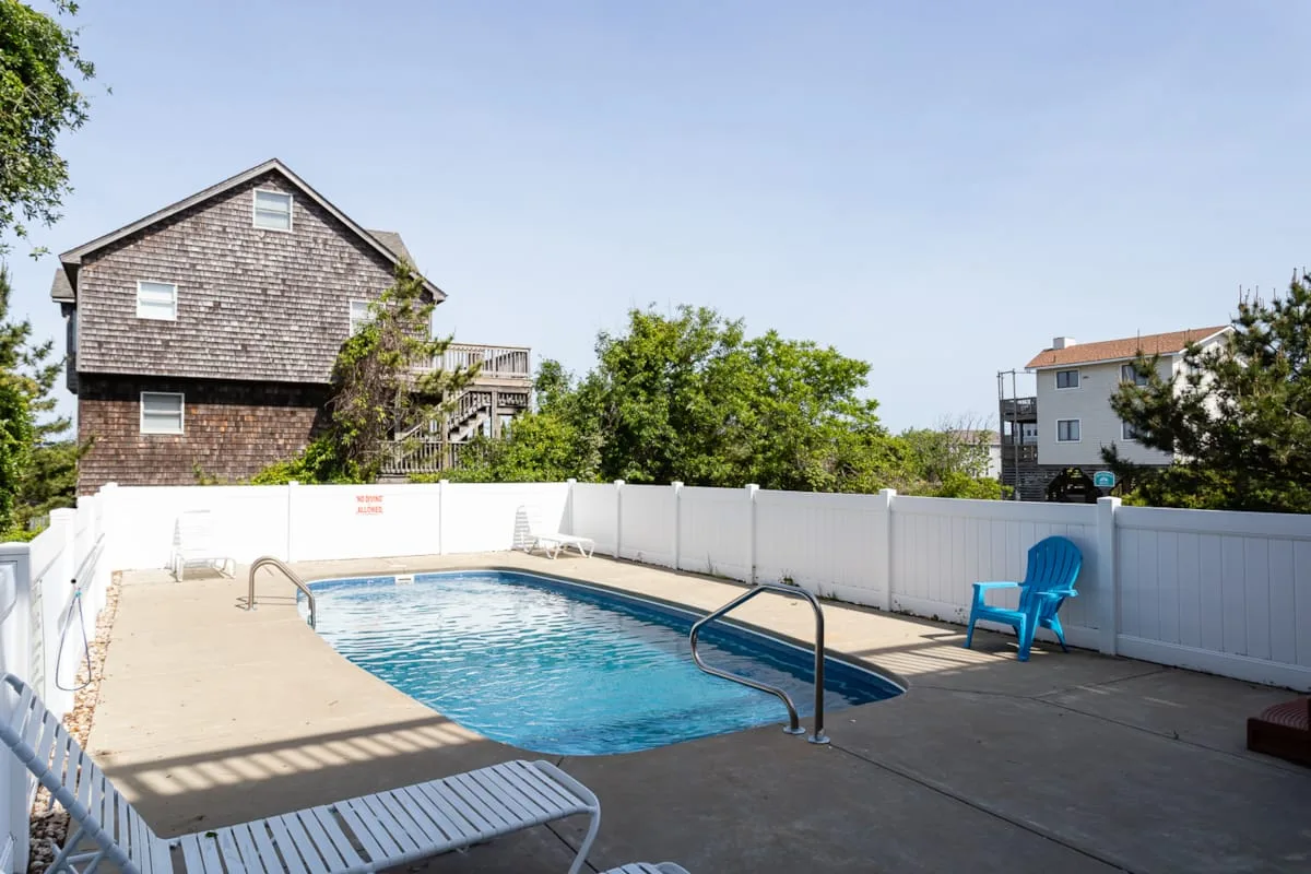 An outdoor swimming pool sits beside a white privacy fence and nearby buildings.