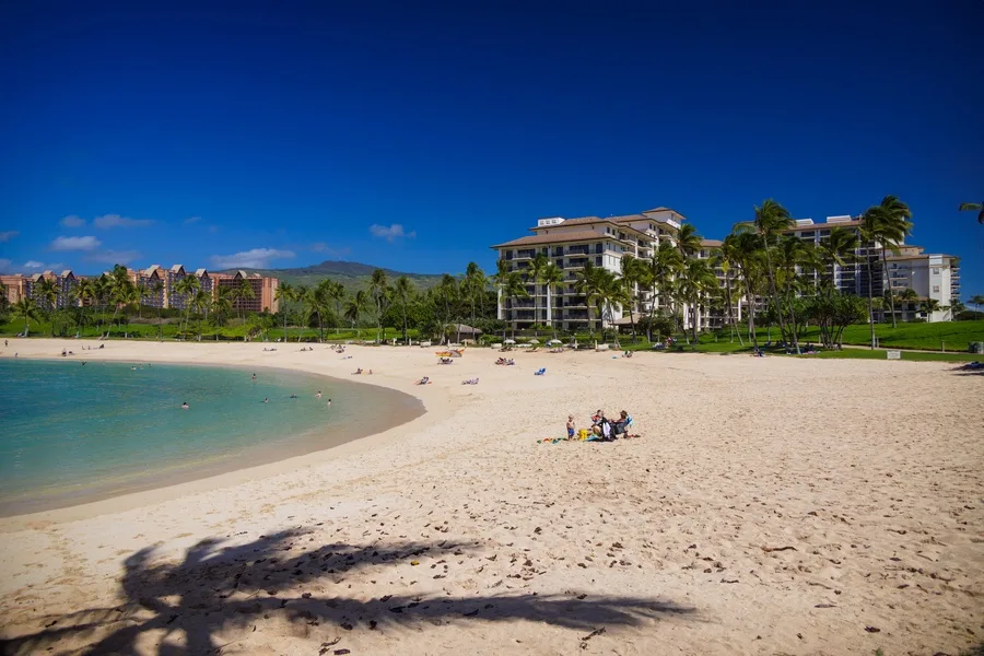 Una playa de arena rodea aguas turquesas tranquilas, con edificios turísticos al fondo.