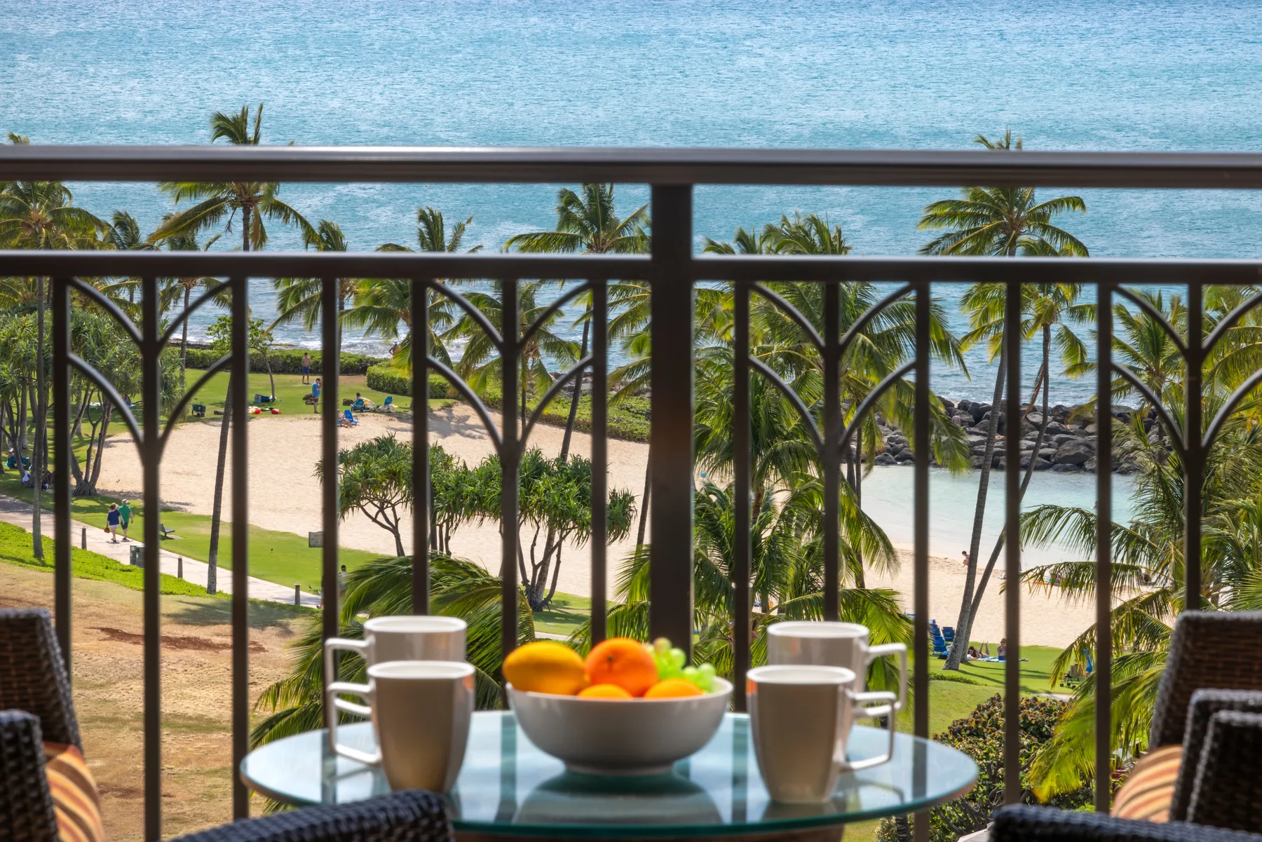 Una mesa de balcón con tazas y fruta da a una playa bordeada de palmeras y al océano.