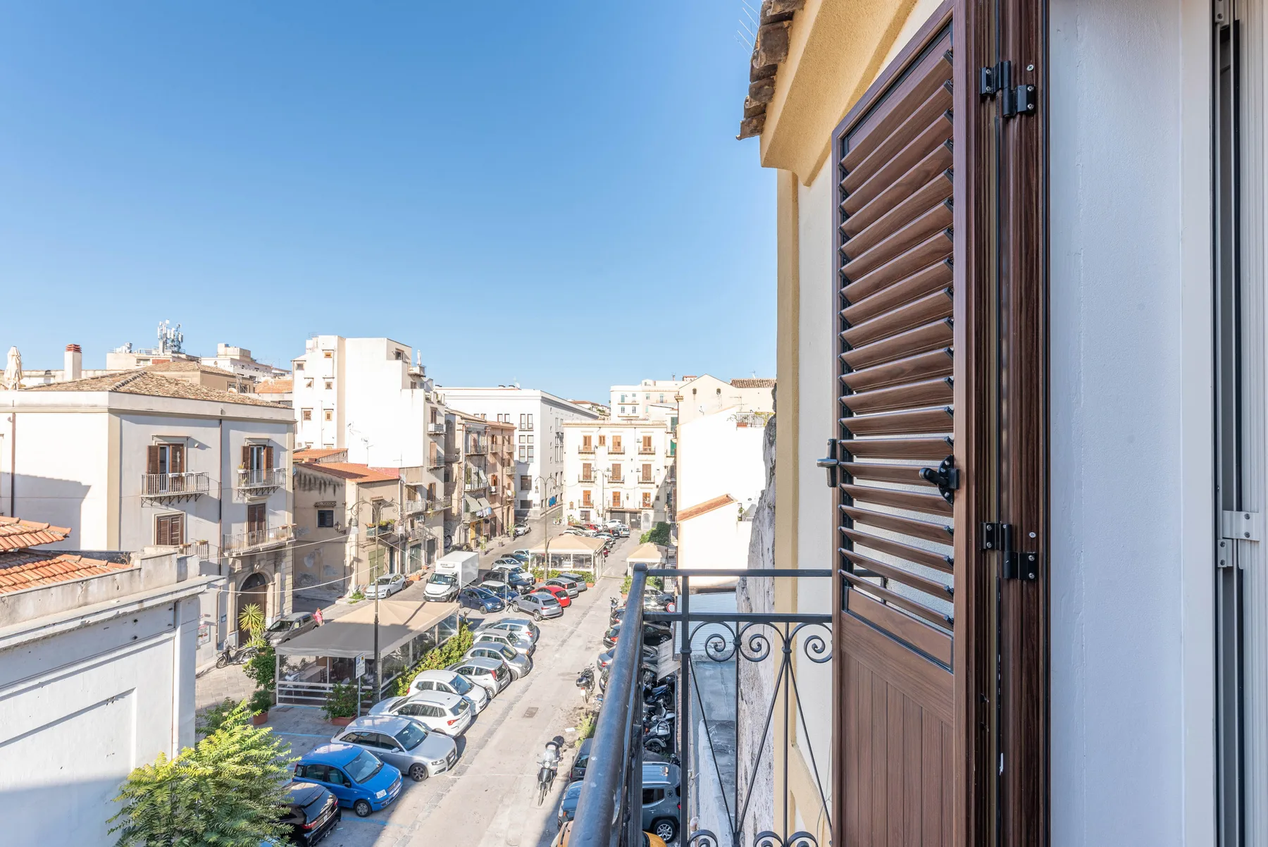 Un balcón da a una calle urbana soleada, bordeada de edificios y coches estacionados.