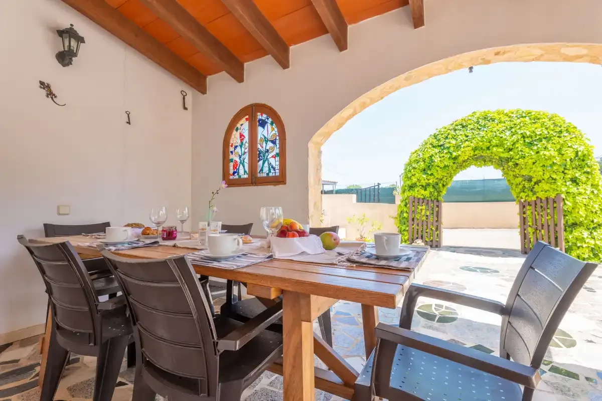 A covered outdoor dining area with a long table and chairs beside an arched garden opening.