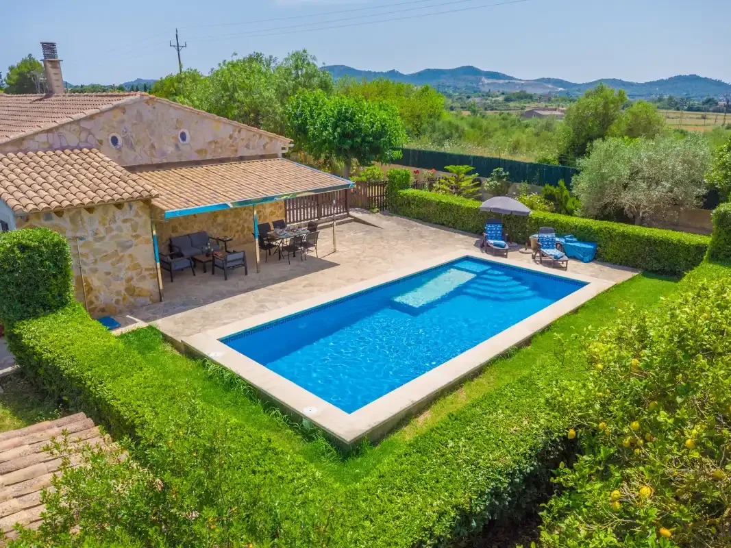 A swimming pool and patio sit beside a stone house, surrounded by greenery.
