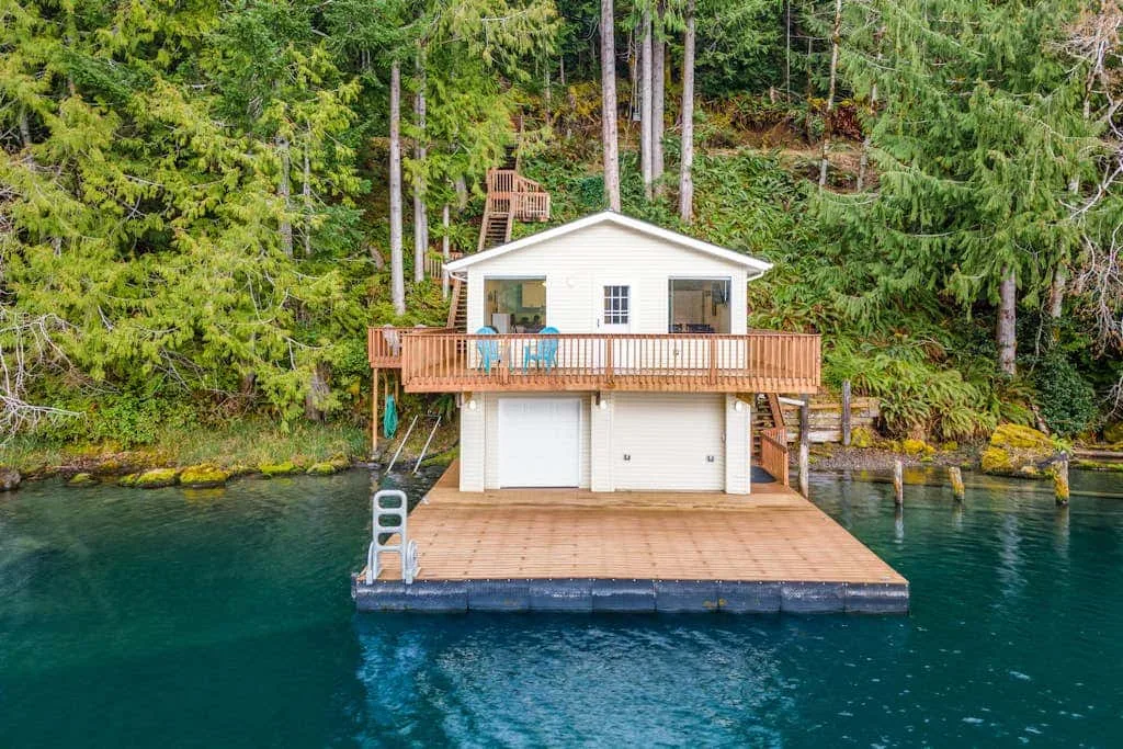 A small waterfront cabin with a wooden deck sits among trees.