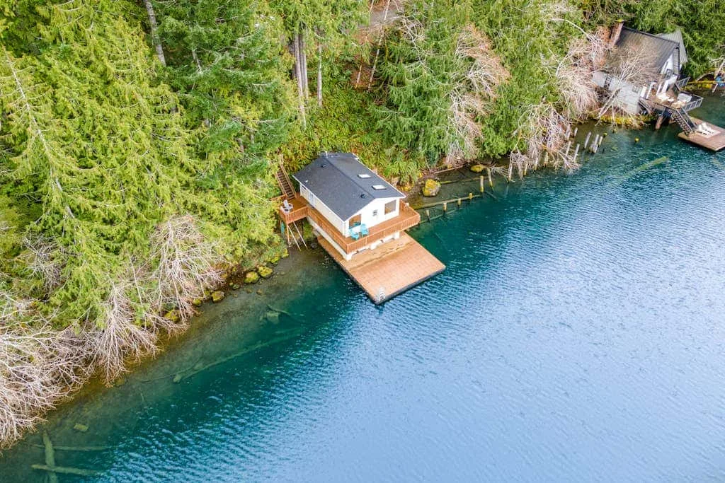 An aerial view shows a small lakeside cabin beside a wooden dock and forested shoreline.