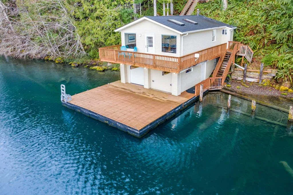 A waterfront house with wooden decks stands beside clear blue-green water.