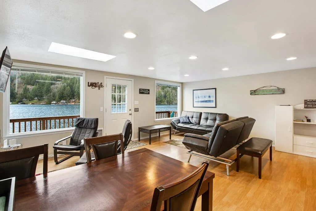 A living and dining room with seating beside large windows overlooking the water.