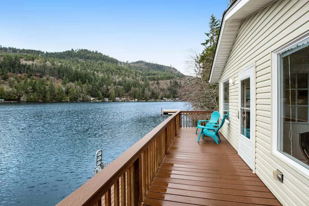 A wooden deck beside a lake has turquoise seating and views of wooded hills.