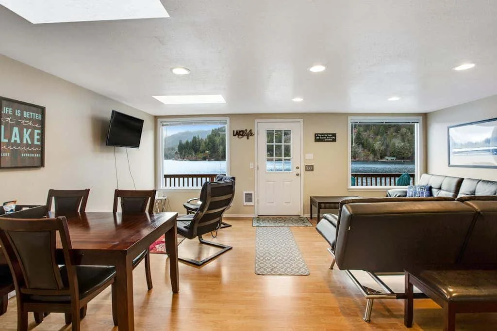 A bright living room with seating, a dining table, and windows overlooking a lake.