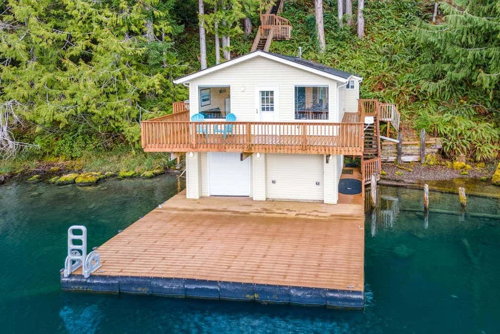 A white lakeside cabin stands above a broad wooden dock surrounded by forest.