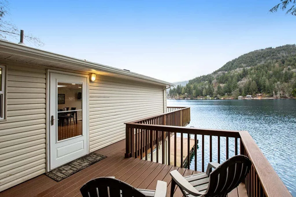 A lakeside deck with seating overlooks the water and a wooded hillside.
