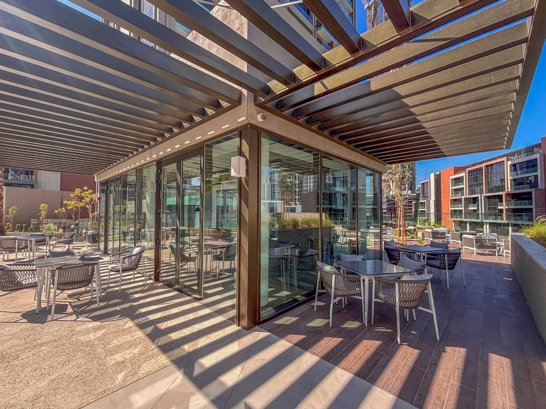 Una zona de comedor exterior acristalada con mesas y asientos bajo una pérgola de lamas.