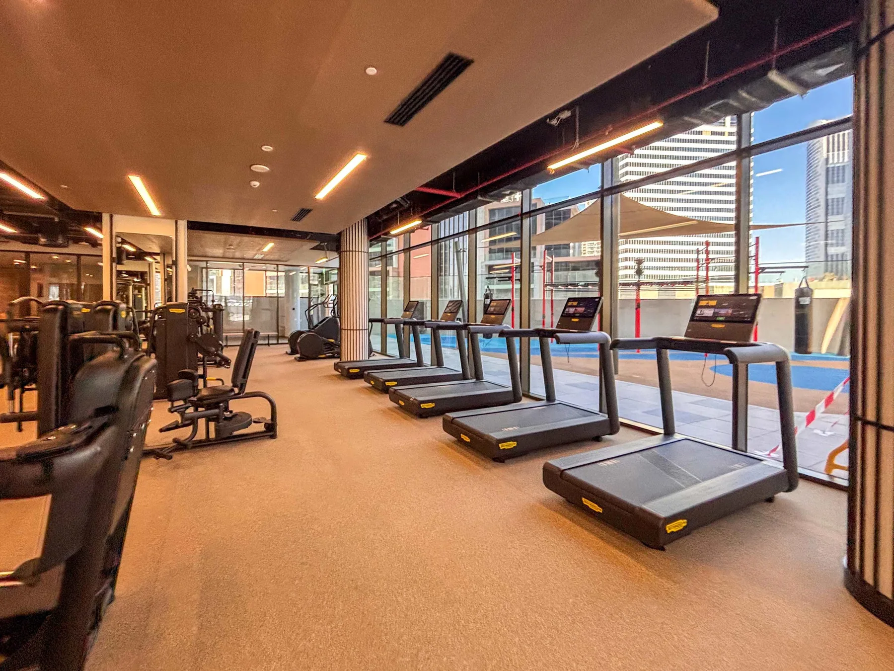 Un gimnasio con cintas de correr junto a grandes ventanas y otros equipos de ejercicio.