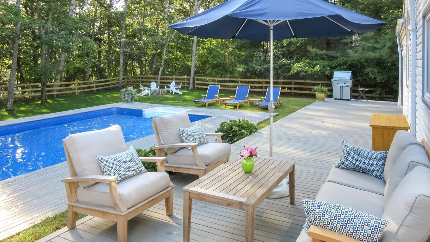 Un patio exterior junto a una piscina, con asientos acolchados bajo una sombrilla grande.