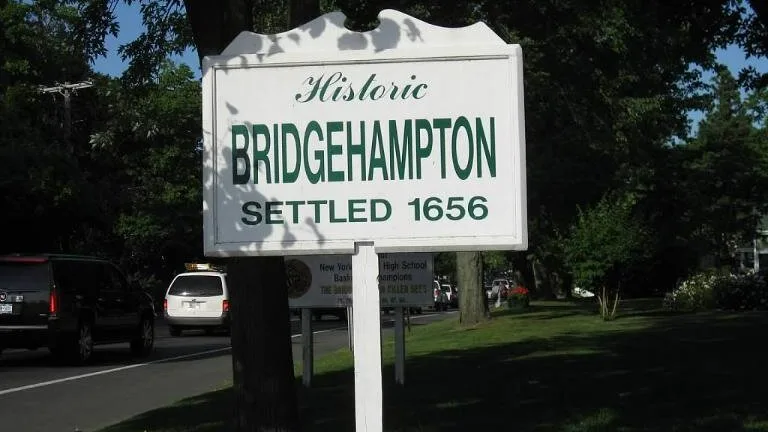 Un letrero junto a la carretera señala el asentamiento histórico de Bridgehampton, con árboles y vehículos estacionados detrás.