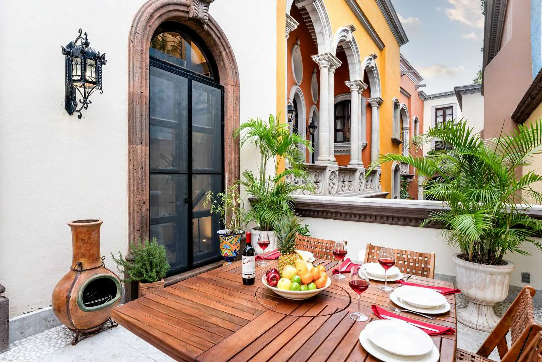 An outdoor dining terrace with a wooden table and potted plants beside colorful buildings.