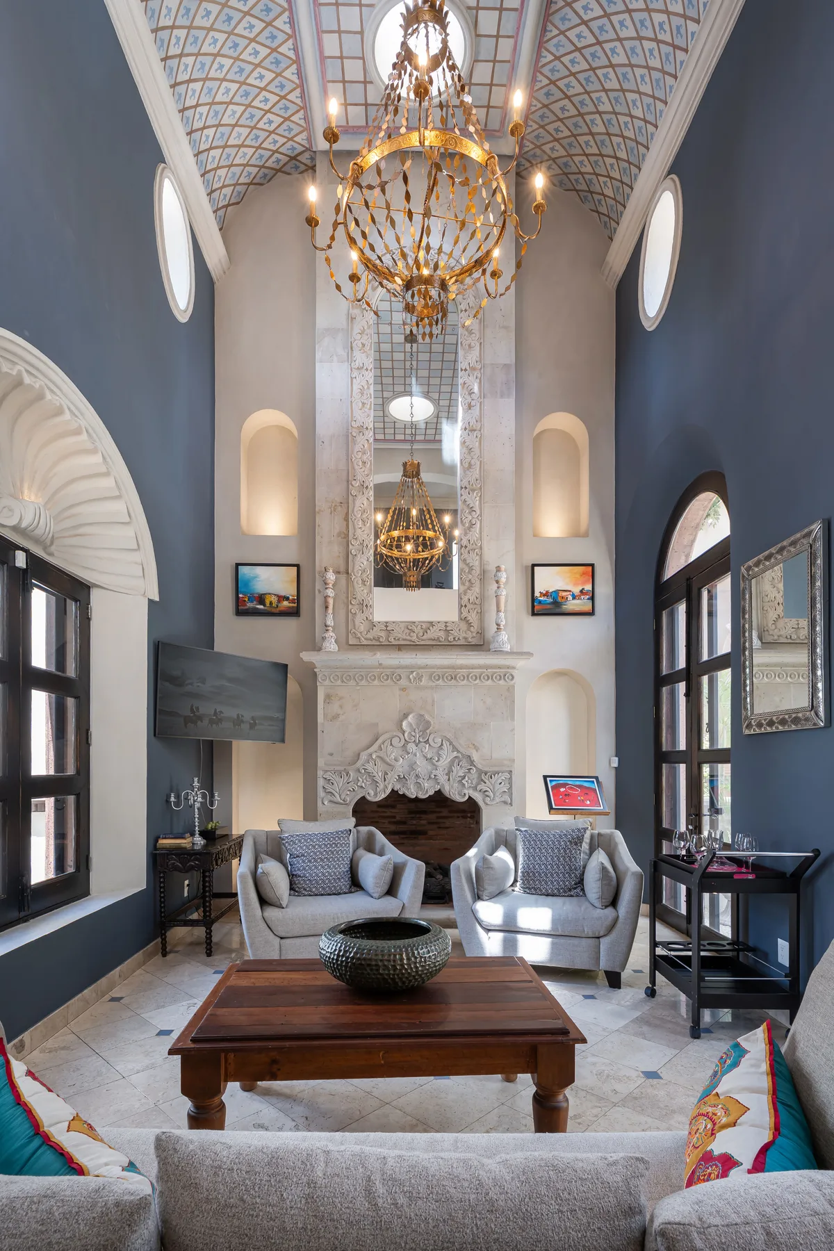 A tall living room features a fireplace, seating, and a large chandelier.