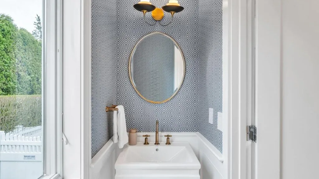 A bathroom vanity with an oval mirror, patterned walls, and a view through the window.