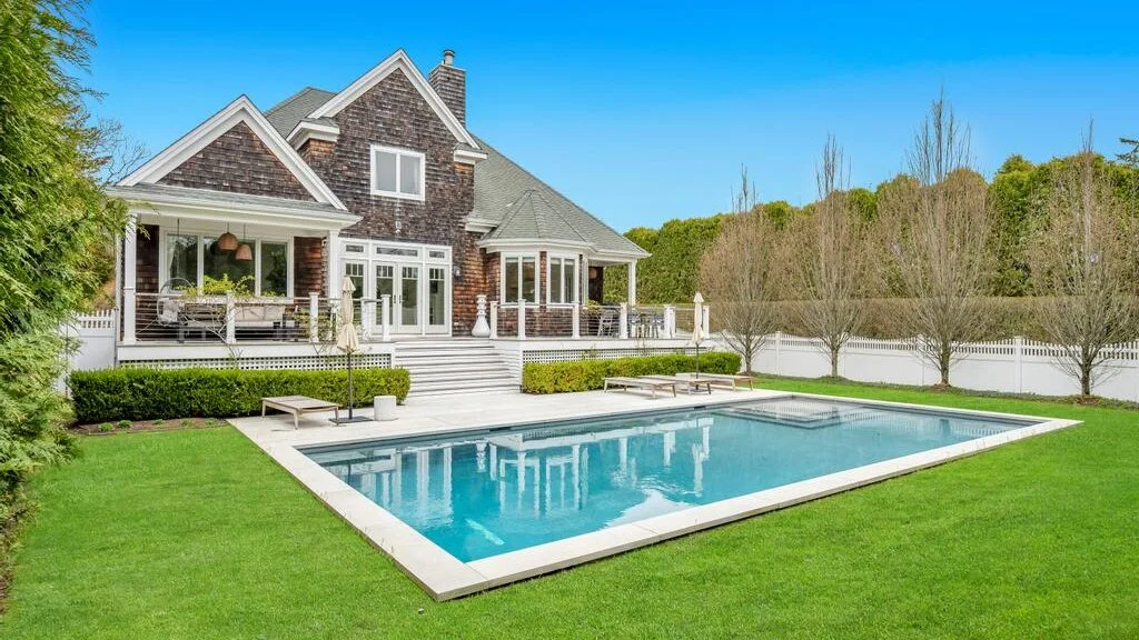 A large house stands beside a rectangular swimming pool and green lawn.