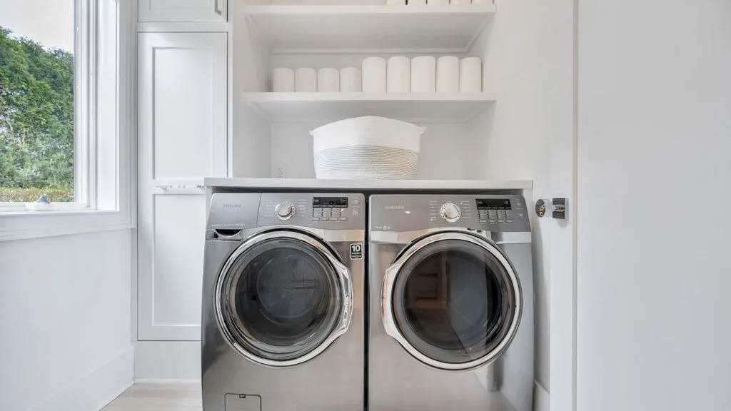 A bright laundry room with a front-loading washer and dryer beneath open shelves.