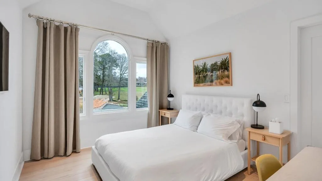 A bright bedroom with a bed, arched window, and beige curtains.