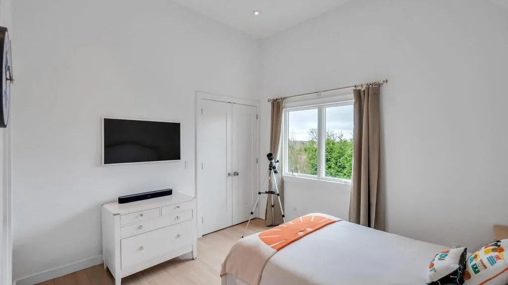 A bright bedroom with a bed, dresser, and a window overlooking greenery.