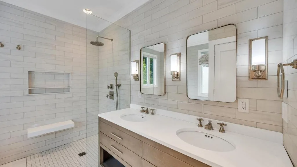 A bright tiled bathroom with a glass shower and a double-sink vanity.