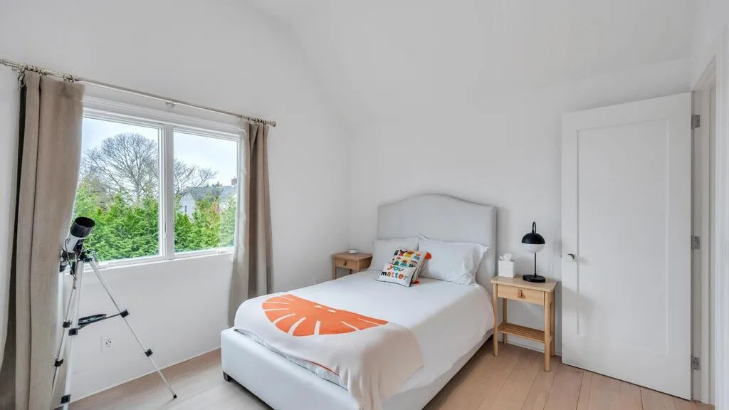 A bright bedroom with a white bed and a window overlooking greenery.