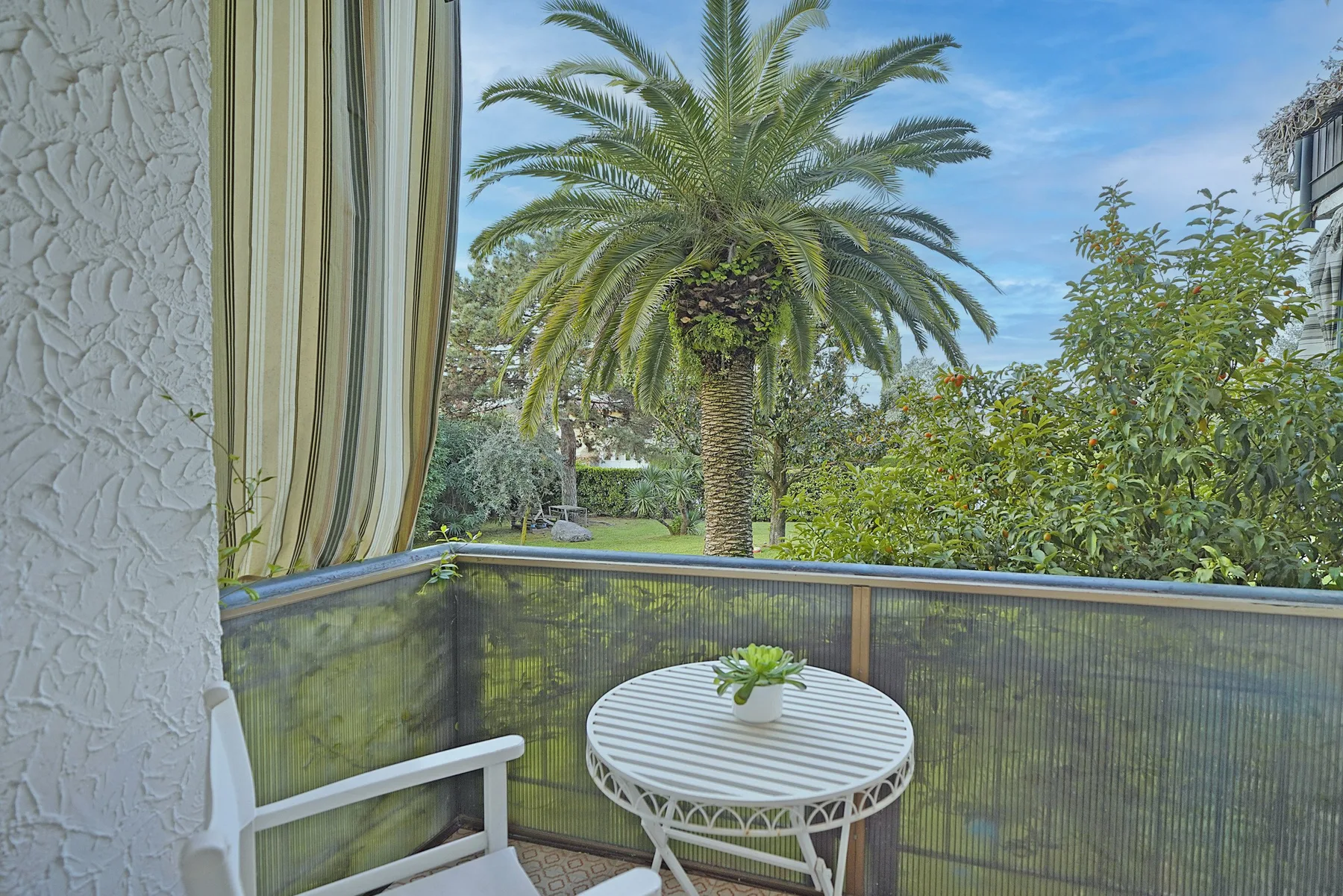 Un pequeño balcón con una mesa redonda da a una palmera y vegetación.