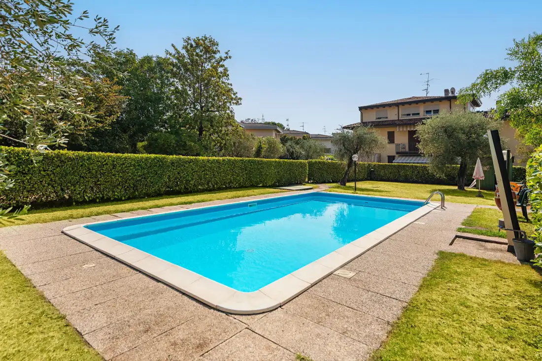 Una piscina al aire libre está rodeada de pavimento, setos y árboles.