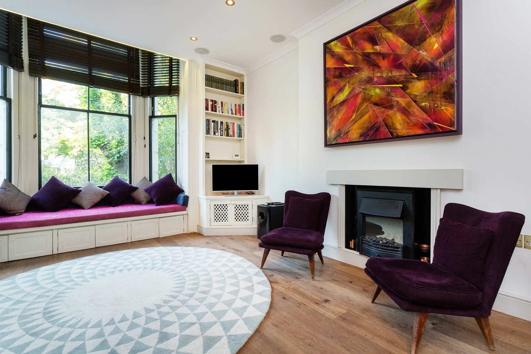 Una sala luminosa con un asiento junto a la ventana y arte colorido sobre la chimenea.