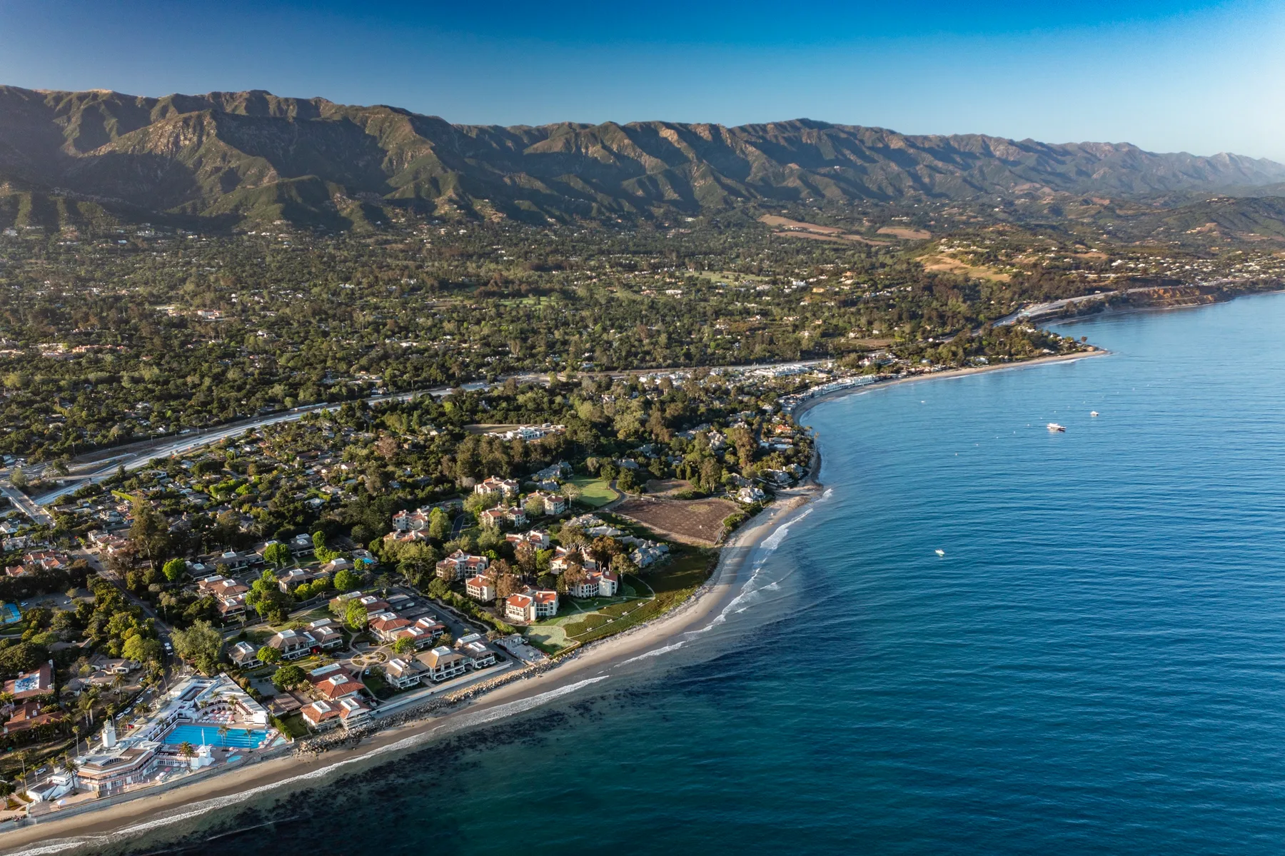 Aerial view of a coastal neighborhood with a curving shoreline and mountains beyond.