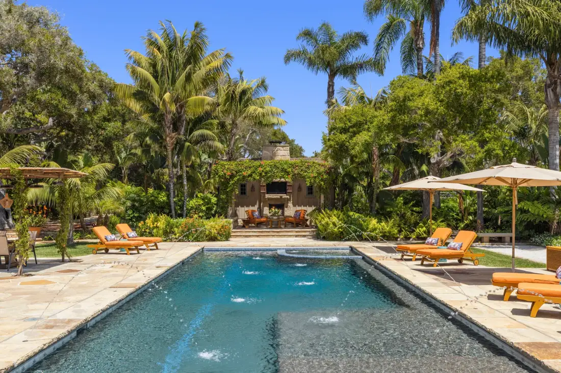A swimming pool sits among palm trees, loungers, and shaded seating.