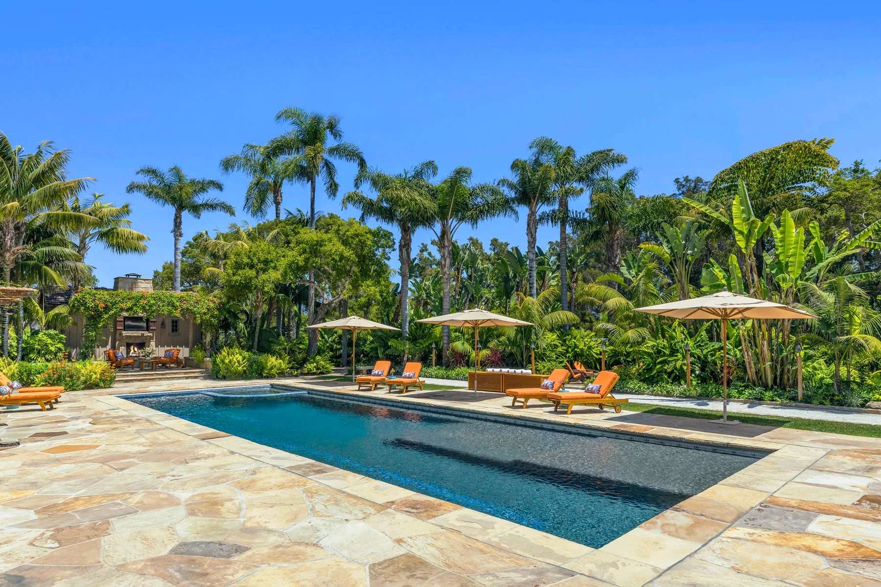 A swimming pool sits beside lounge seating and umbrellas in a tropical garden.