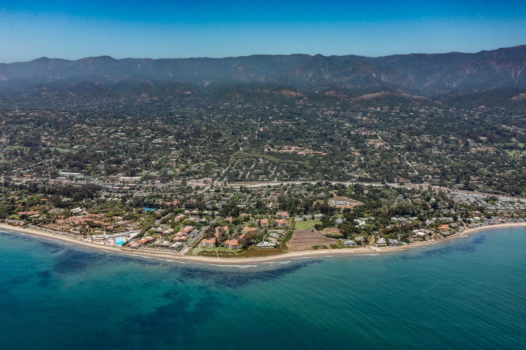 An aerial view of a coastal town along a curving shoreline, with mountains in the distance.