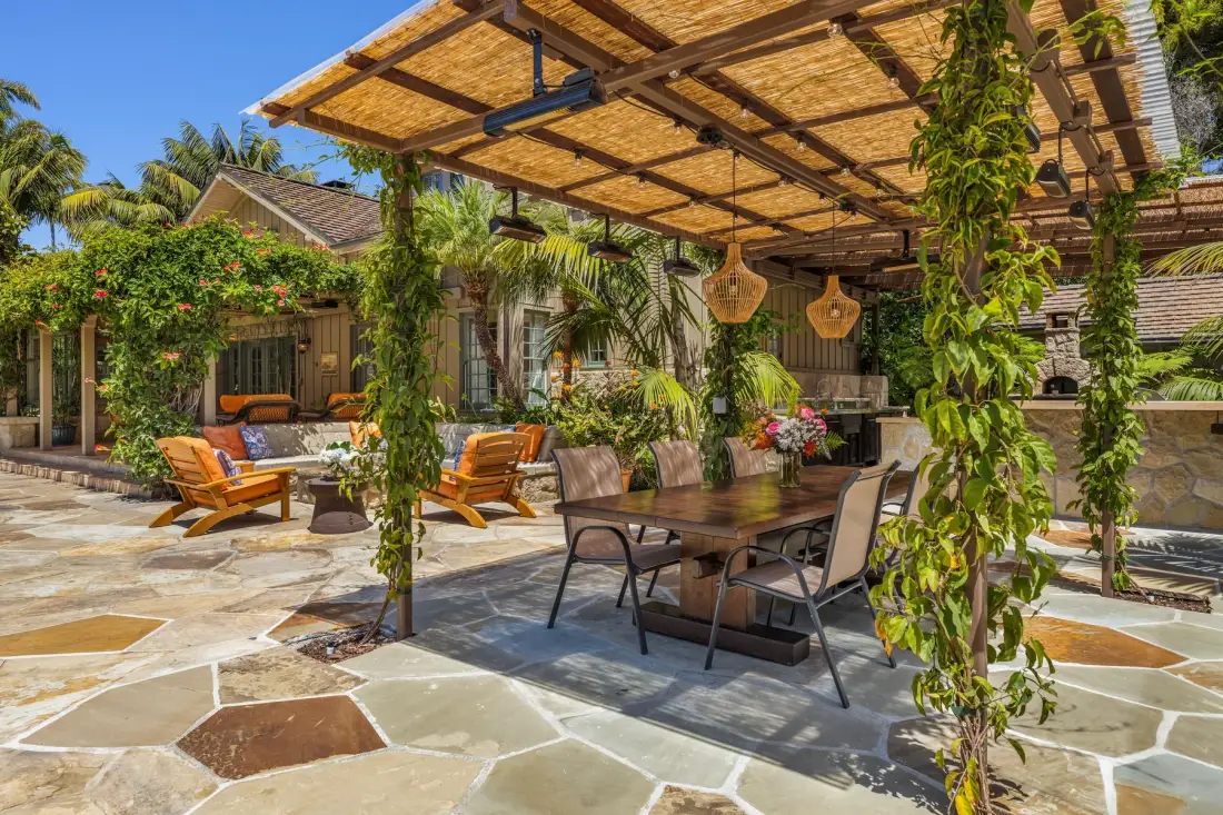 An outdoor dining area sits beneath a wooden pergola, surrounded by greenery.